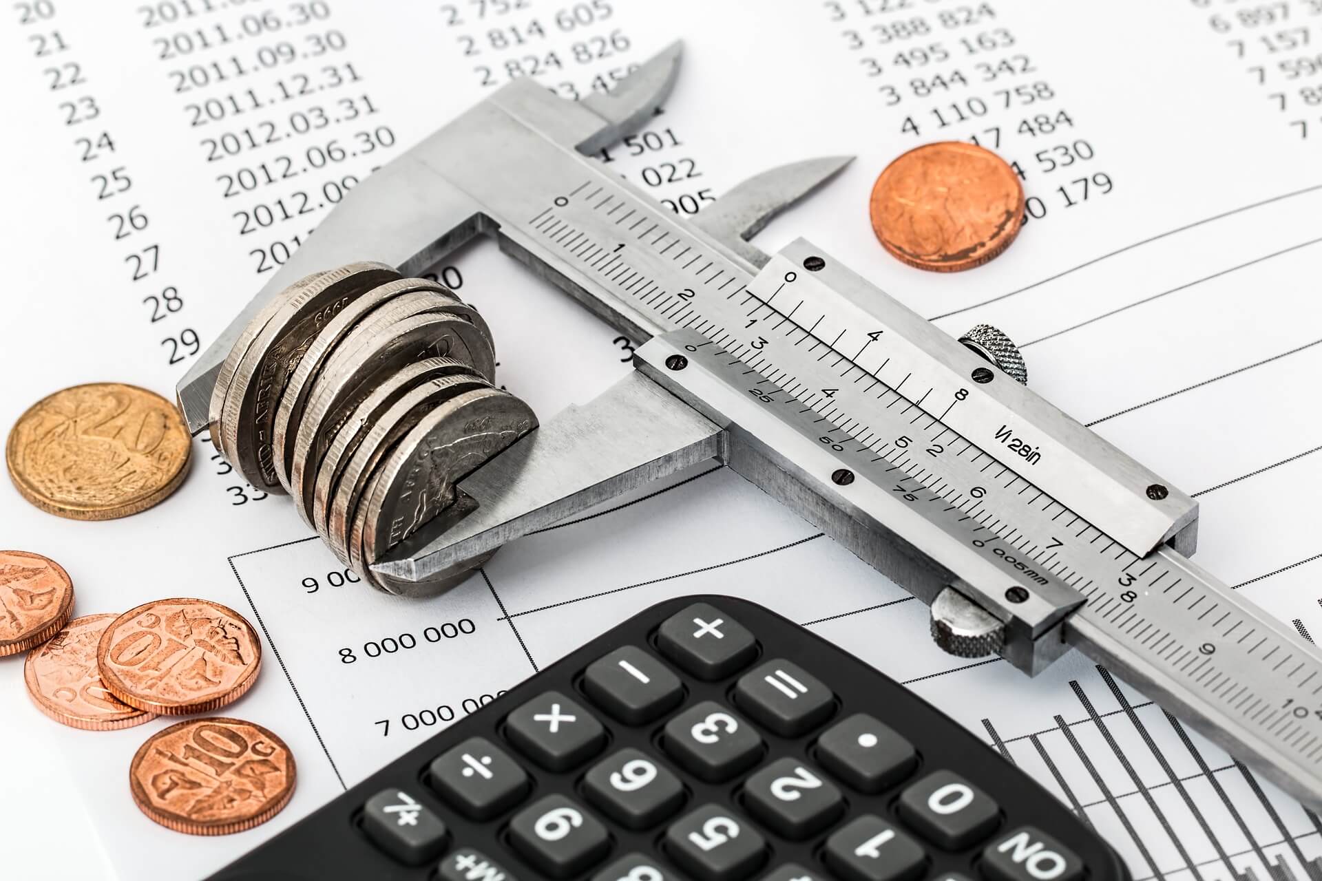 A pair of metal calipers holding a stack of silver coins, lying atop a spreadsheet next to a calculator and some copper coins.