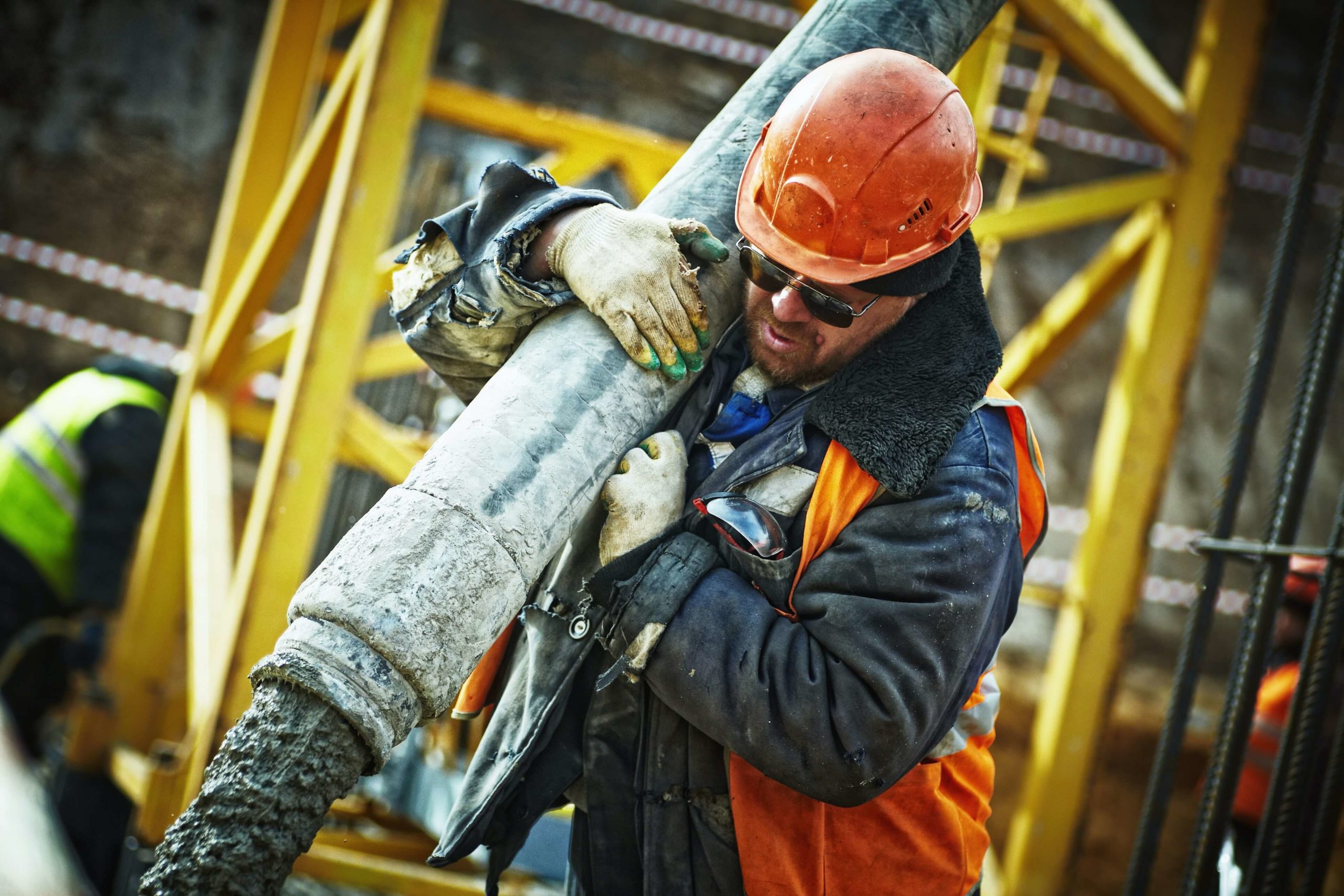 A man in a hardhat and vest holding a pipe emitting liquid concrete on a construction site.