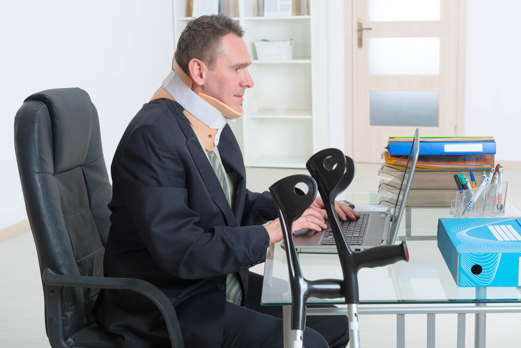 An individual wearing a neck brace and a dark suit is sitting at a desk working on a laptop, with a pair of crutches leaning against the desk nearby.