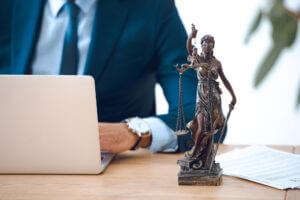 View of the back of a laptop while a lawyer dressed in a suit works at a desk, with a desktop statue of Lady Justice sitting to the side.