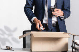 Medium shot of an individual in a business suit standing at a desk, placing personal items into a cardboard box as they prepare to leave the office after being terminated.