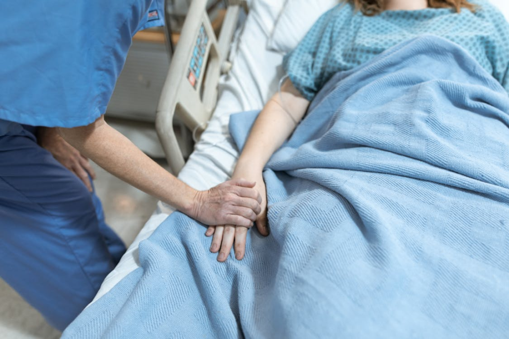 A medical professional wearing scrubs holds a patient's hand lying in a medical bed, assuring them.
