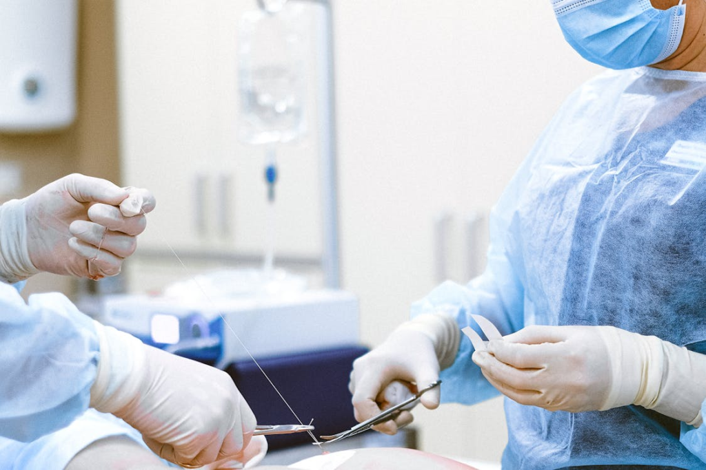 Two medical professionals wearing plastic gloves, surgical gowns, and masks, cut off a piece of suture thread.